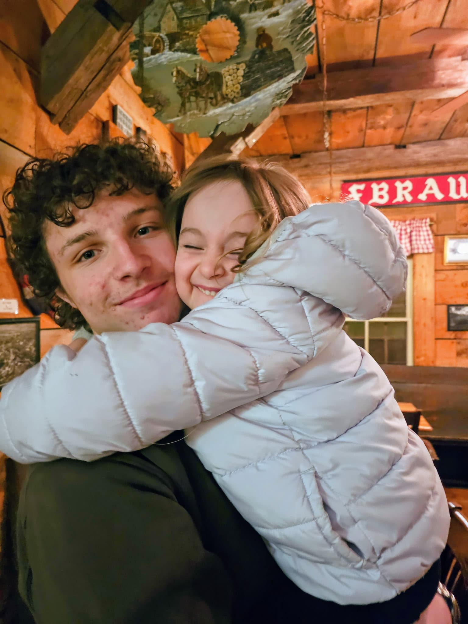 A curly-haired young man smiles while holding a young girl in a white puffer jacket.