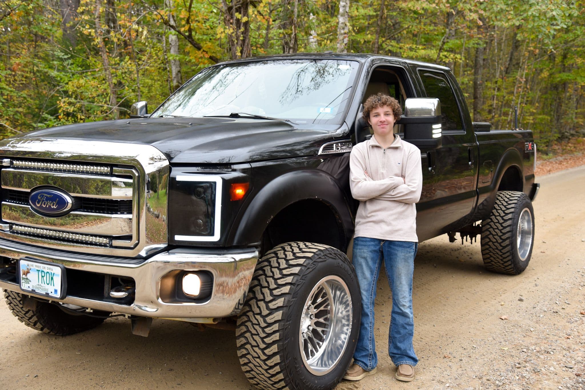 Curly-haired teen stands with arms crossed next to a lifted black Ford F-250 truck.